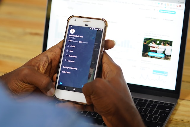 صورة لشخص يحمل هاتفا أمام جهاز كمبيوتر محمول يشاهد القائمة الجانبية لتويتر مع خيار الإعدادات والخصوصية.