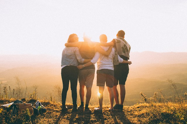 Mehrere Freunde halten sich gegenseitig in den Armen, stehen am Rande eines Hügels und betrachten den Sonnenuntergang.