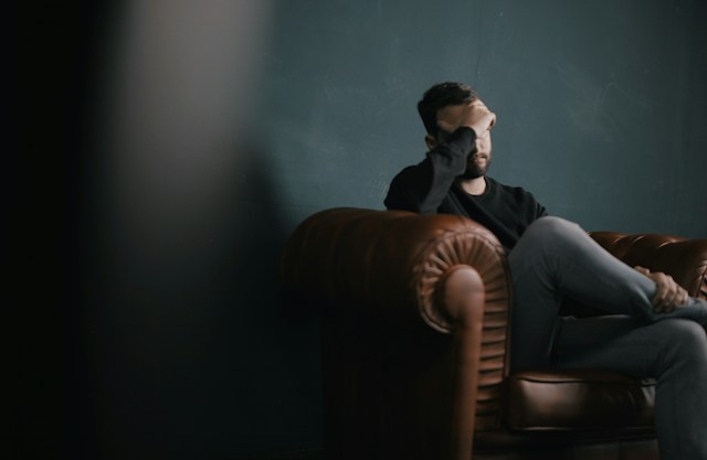 Eine Person, die im Schneidersitz auf einer braunen Couch sitzt und sich mit einer Hand den Kopf hält.