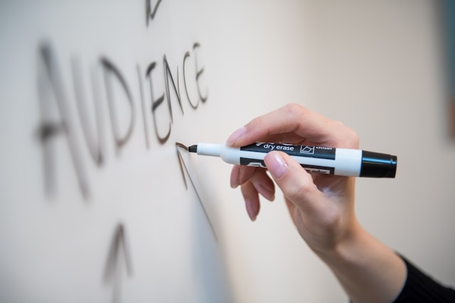 Eine Person schreibt das Wort Publikum mit einem schwarzen Stift auf eine Tafel.