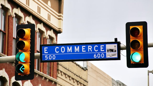 Ein blaues Straßenschild mit "E Commerce" zwischen zwei Ampeln mit grünem Licht.
