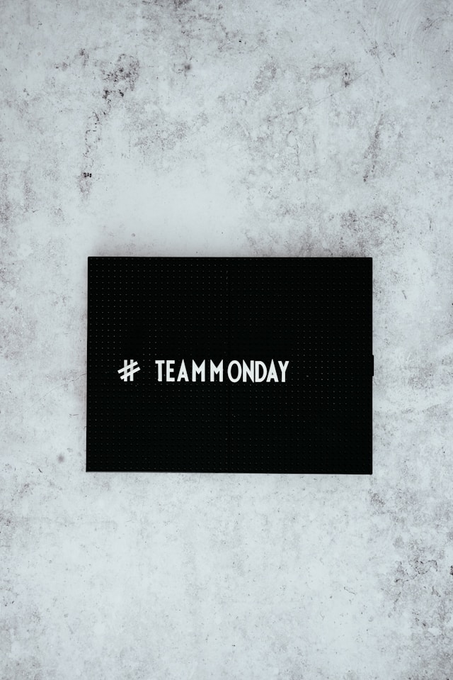 Una pizarra con el hashtag "#teammonday" contra una pared gris.
