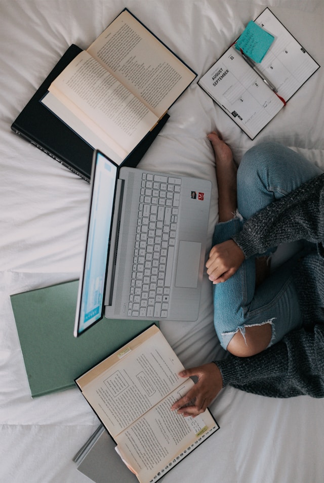 Una persona se sienta en una cama blanca con un portátil gris y consulta un libro con notas que ha escrito.

