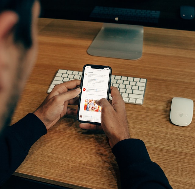 Une photo d'un homme appuyé sur une plate-forme en bois tout en regardant un X ou une timeline Twitter sur un smartphone noir.