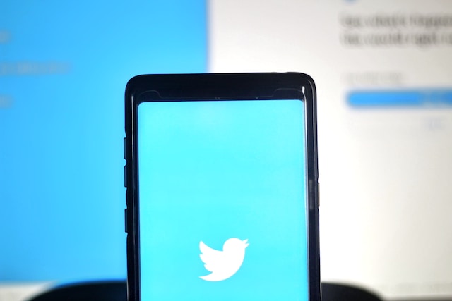 Gambar ponsel Samsung berwarna hitam dengan layar biru Twitter terbuka dengan latar belakang biru dan putih yang kabur. 
