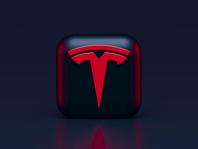 Render 3D logo Tesla berwarna merah di atas kubus hitam.
