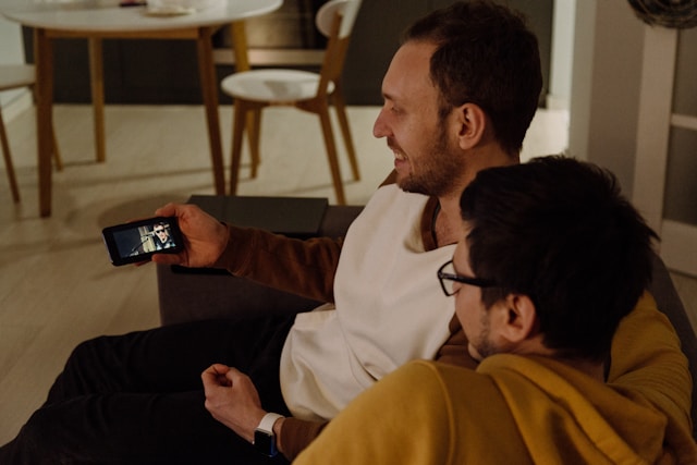 Due uomini siedono uno accanto all'altro su un divano e guardano un video su uno smartphone.

