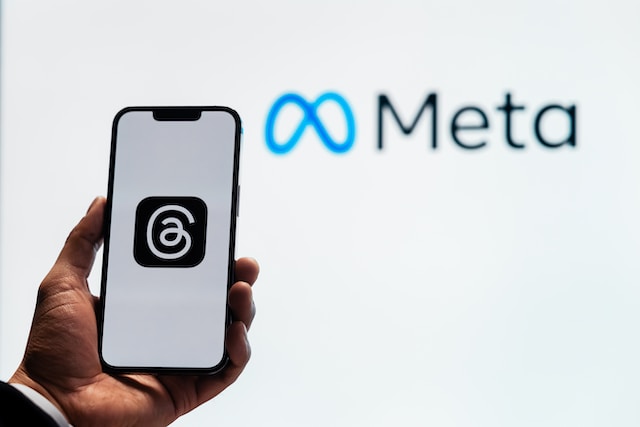 巨大なMetaのロゴを背景に、Threadsのロゴが表示された携帯電話を持つ人の写真。