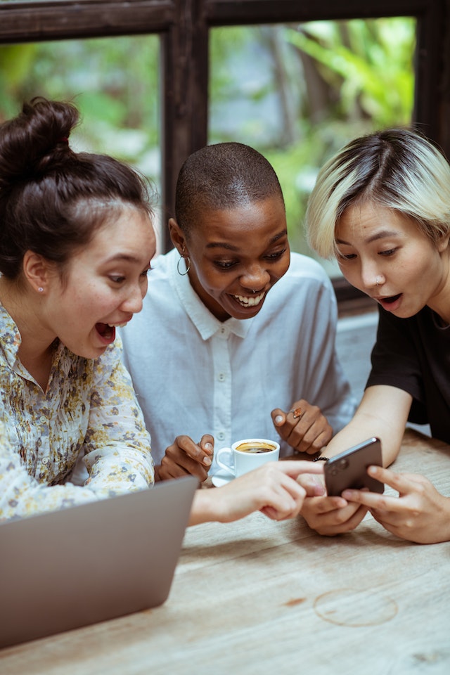 Três mulheres olham com entusiasmo para um smartphone. 