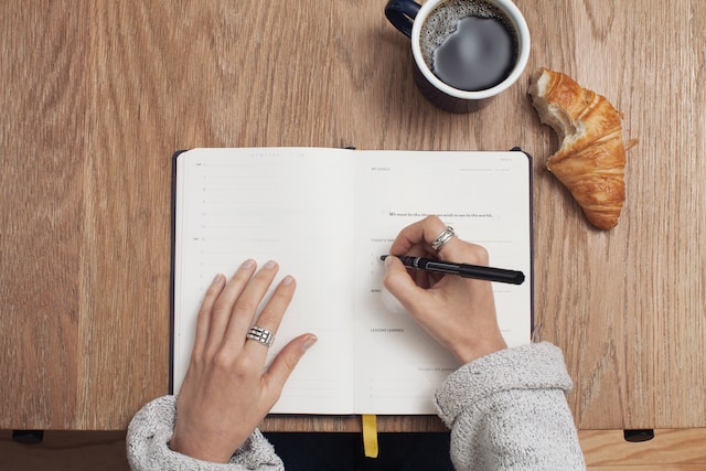 Uma fotografia de alguém a escrever num livro com uma chávena de chá e uma bolacha ao lado.