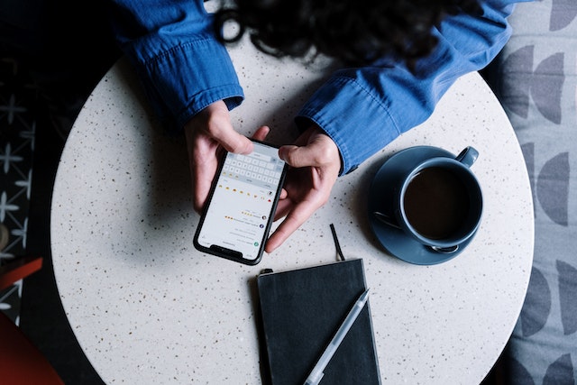 A photograph of someone in a blue denim jacket using their phone at a table with a writing pad and teacup.