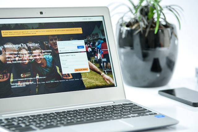 A photograph of a gray laptop on a table next to a vase displaying Twitter’s welcome page.
