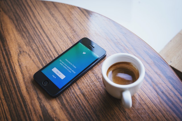 An iPhone with the Twitter app open, placed on a wooden table next to a cup of coffee.