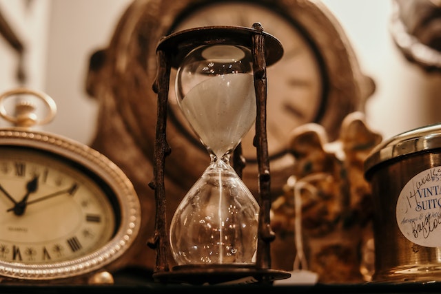 A classic hourglass placed next to two clocks. One clock is beside it, and the other is behind it.