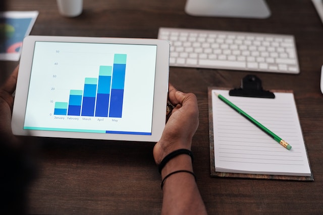 A picture of a person holding a tablet while working on a desk with a bar chart on the screen.