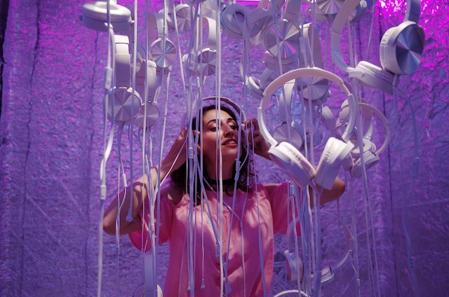 A woman wearing white headphones while standing among other white headphones and cables dangling from the ceiling.