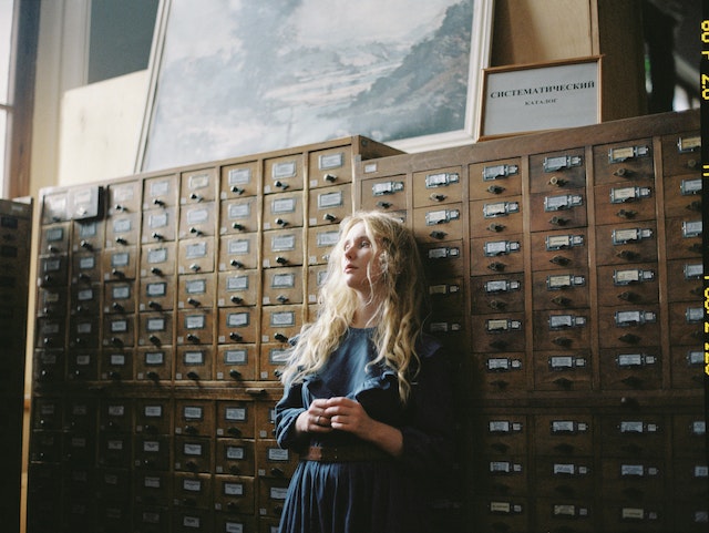 A picture of a woman leaning against a physical archive of tiny labeled shelves.