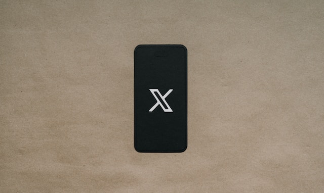 A picture of the X logo displayed on a black smartphone placed on brown paper.