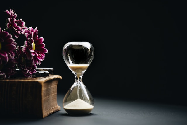 A picture of a clear hourglass placed on a dark background with a pink flower beside it. 