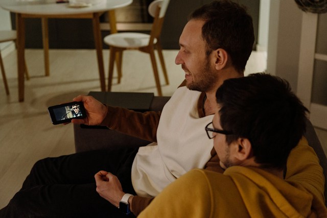 Two men watch a video on a smartphone while sitting on a couch.