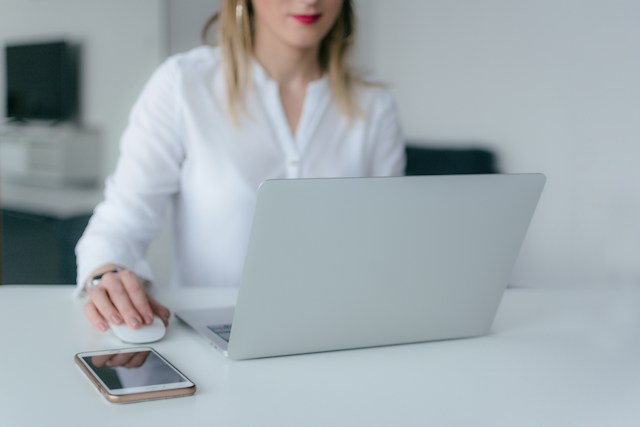 A woman smiles as she uses a Bluetooth mouse to work on her laptop and has her cell phone beside it on the desk.