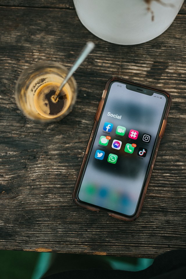 A folder containing various social media apps on an iPhone placed on a wooden table beside a cup of coffee.
