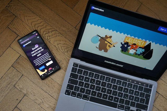 Mastodon on an Android smartphone next to a gray Macbook Pro with the Mastodon website on its display. 

