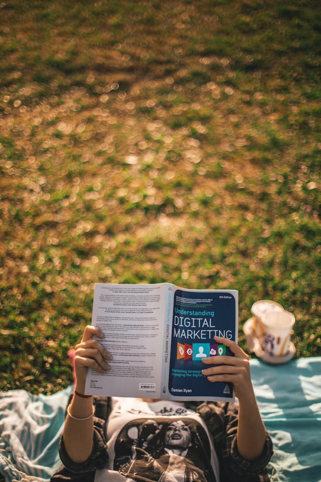 A person lies down on the grass and reads a book about digital marketing.