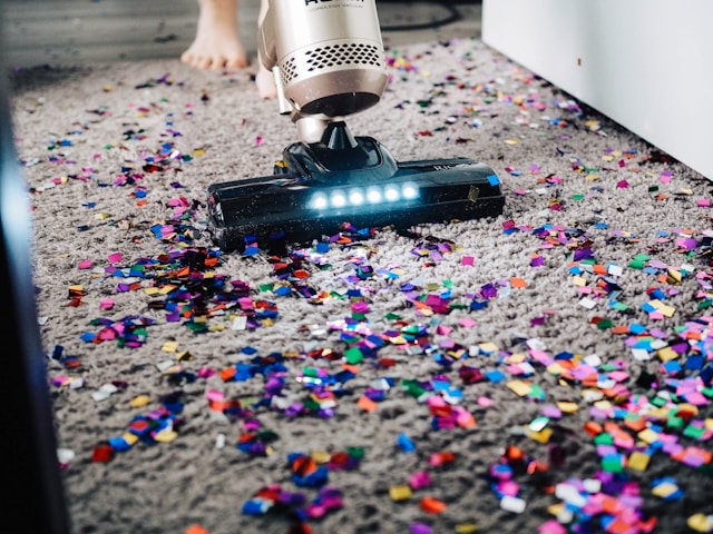 A person uses a vacuum cleaner to remove confetti from their gray carpet.
