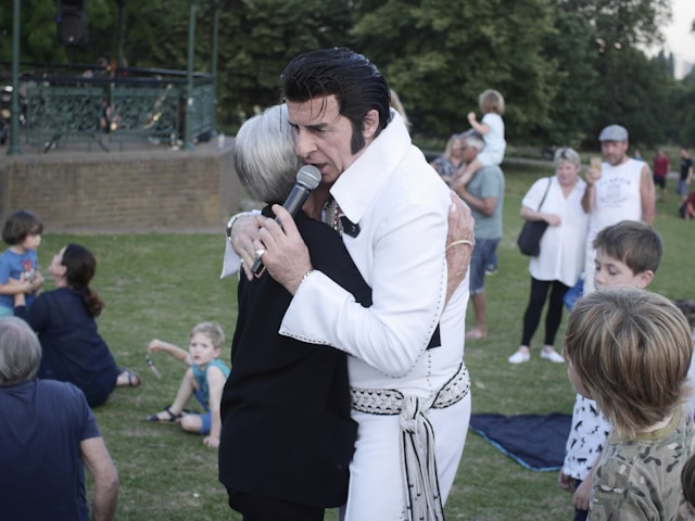 An Elvis Presley impersonator with white clothes holds a microphone and hugs a woman.