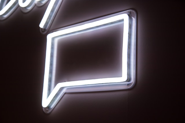 A white neon sign of a square chat bubble on a wall.
