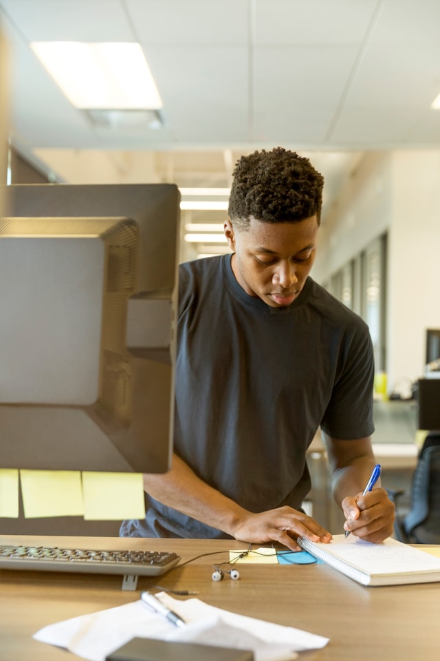 A man in a black shirt writes a note in a book with a blue pen.
