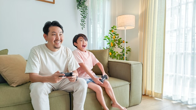 A man and a child sit on a couch and play a game with black wireless controllers.

