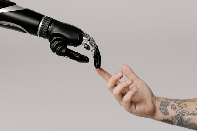 A black bionic hand’s finger touches a tattooed human’s index finger.
