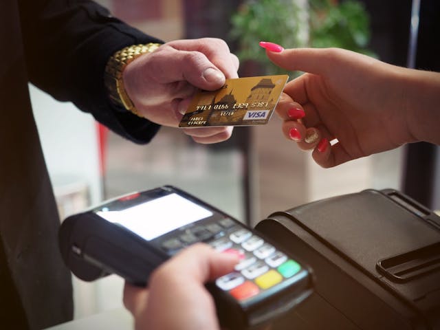 An individual with a black coat and a gold watch hands over a gold debit card to another person.
