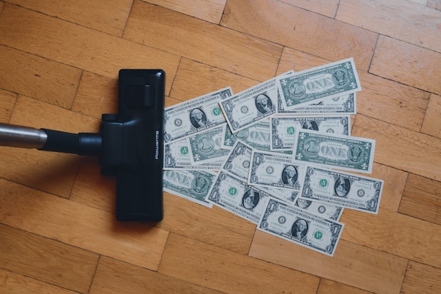 A black vacuum cleaner over several one-dollar bills on a wooden floor.