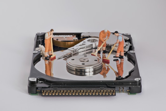 Three figurines with orange overalls brush the drive platter of a hard disk without its outer case.