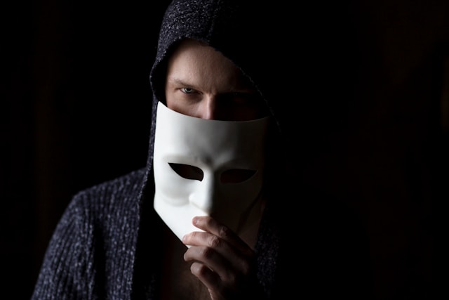 A man with a hoodie holds a white mask below their face.
