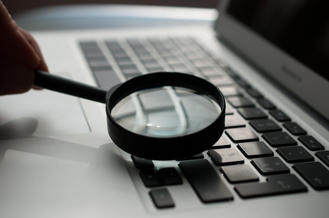 A person holds a black magnifying glass next to a gray MacBook’s keyboard.