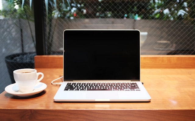 Un Macbook gris junto a una taza blanca sobre una mesa marrón.