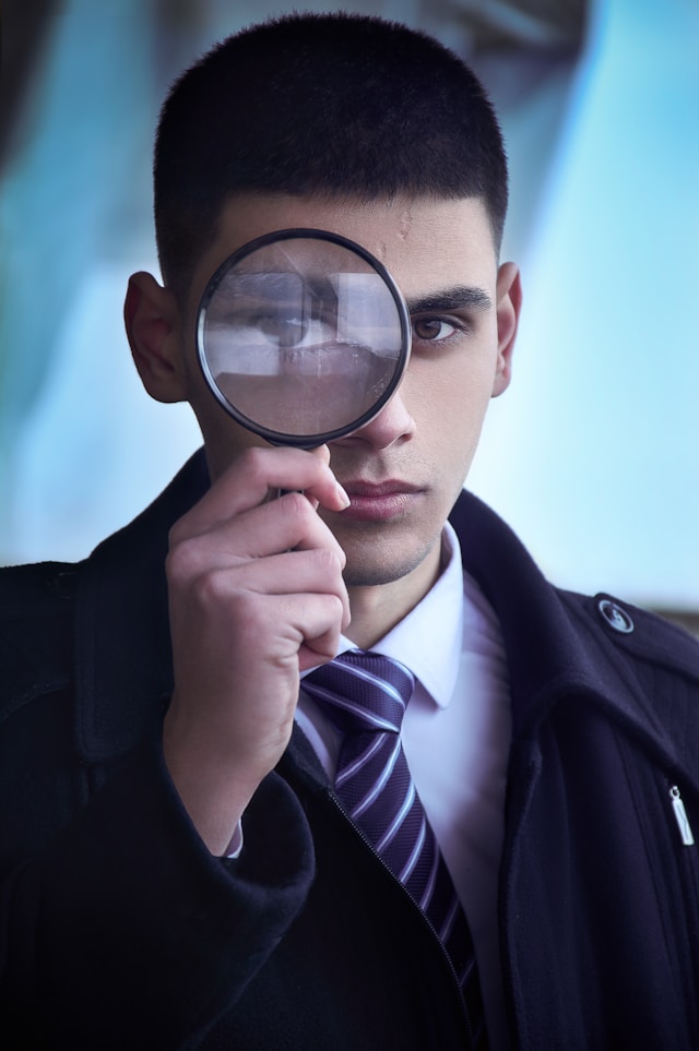 A man with a black suit, pink shirt, and purple tie holds a magnifying glass close to their eye.
