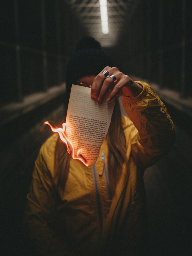 A woman with a yellow jacket and a black beanie holds a piece of paper on fire.
