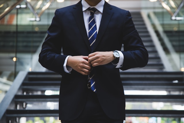 Uma pessoa com um fato azul e uma camisa branca está de pé numa escadaria.
