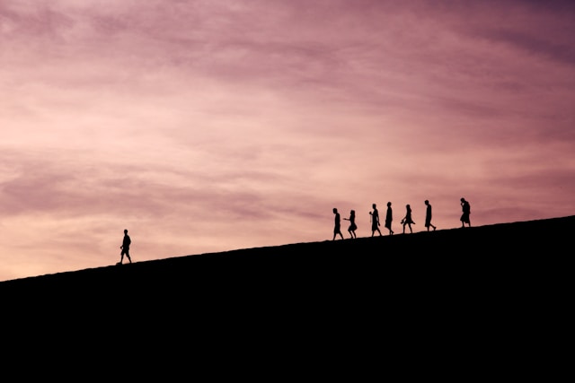 The silhouette of several people on a downward slope against a cloudy sky lit up by sunset.