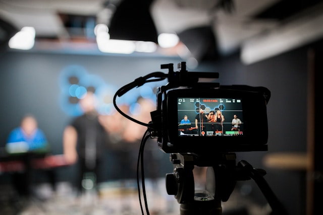 A black DSLR camera on a tripod stand takes a video of a group of people.