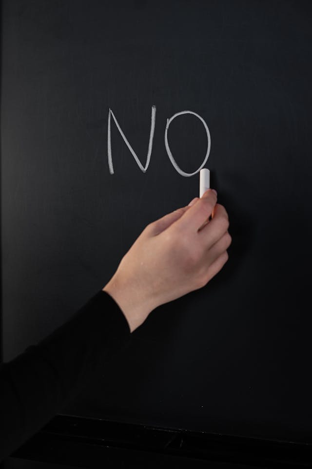 A person holds a piece of white chalk next to the word “no” on a chalkboard.