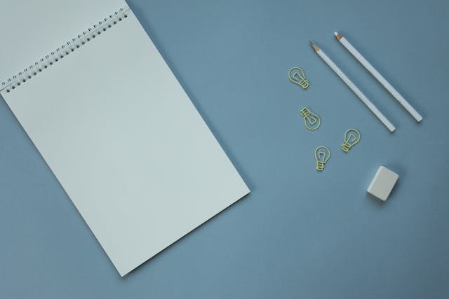 Buku catatan spiral kosong di samping penghapus, dua pensil, dan empat bola lampu miniatur berwarna kuning dengan latar belakang biru muda.