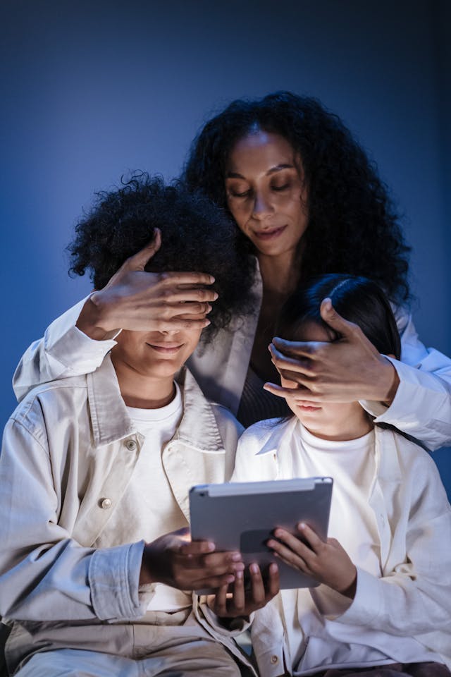 Dua orang anak memegang sebuah tablet berwarna abu-abu, dan seorang wanita menutup mata mereka.