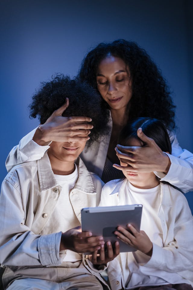 Two children hold a gray tablet, and a woman covers their eyes.