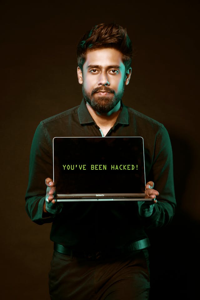 Un bărbat ține în mână un Macbook Pro gri pe al cărui ecran apare textul "You've been hacked".
